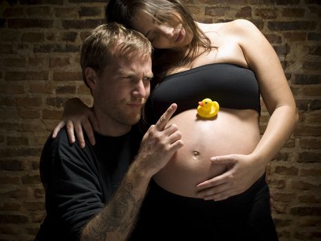A man pointing his finger at the rubber duck over his wife's pregnant belly.