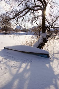 Old wooden boat covered with snow resting near tree on the coast of the river.