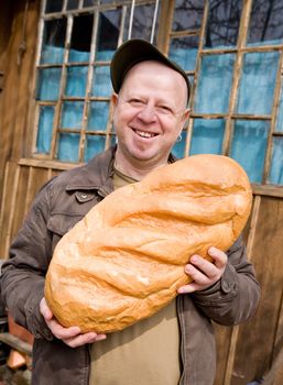 The laughing amusing man holds a huge loaf of bread near the rural house

