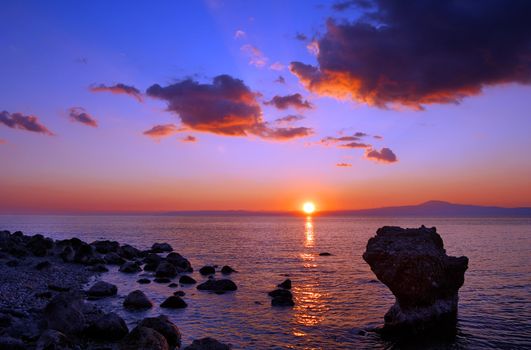 Image shows a sunset over the Messinian bay, Greece, with a rocky seascape in the foreground