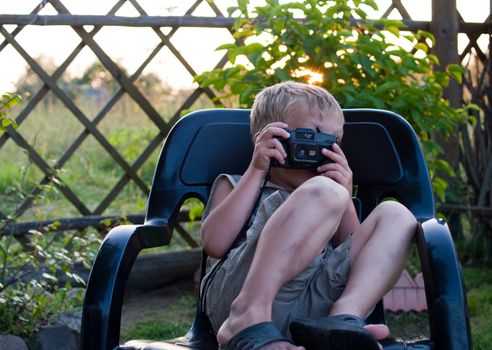 The child with camera sits in a black plastic armchair in the warm summer evening.