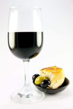 Glass of red wine with three green olives and crusty bread in a small black bowl on a plain background