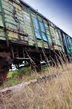A low a angled view of a derelict train carriage in a sideing. Overgrown grass growing to foreground. Train in a derelict condition with rust and peeling green paint.