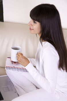 Beautiful latina woman is sitting on a cream color sofa with her computer and a cup of tea working from home