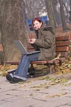 Image of a smiling redheaded girl holding her glasses in her hands and sitting on a bench with a laptop in her lap in an autumn park.Shot with Canon 70-200mm f/2.8L IS USM