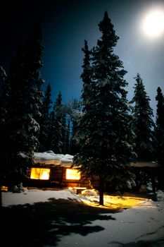 Yukon/Alaska trapline log-cabin fully illuminated at full-moon night in snowy winter.