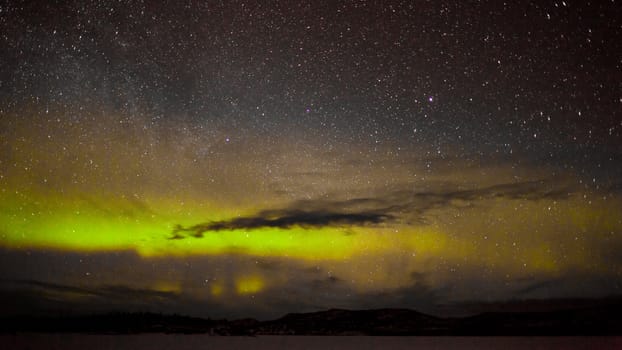 Night sky with myriad of stars with northern lights (aurora borealis) substorm above silhouette of hills and clouds.