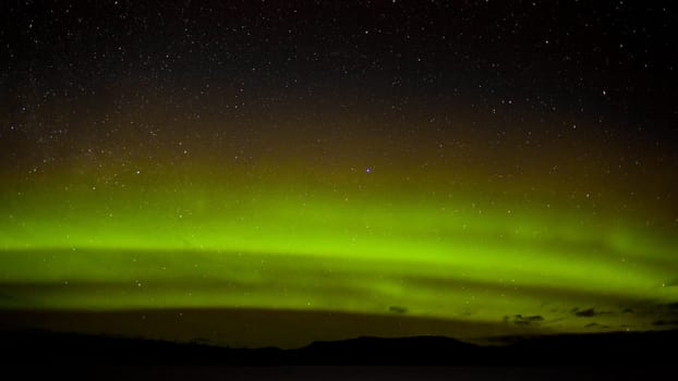 Night sky with myriad of stars with green northern lights (aurora borealis) substorm above silhouette of hills and clouds.
