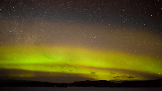 Night sky with myriad of stars with northern lights (aurora borealis) substorm above silhouette of hills and clouds.