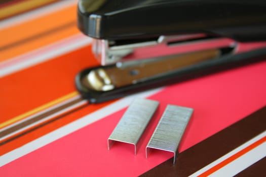 Close up with a shallow DOF of staples and a stapler in the background, all on a very colorful notebook.