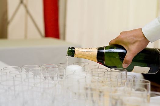The hand of the waiter holds a bottle of champagne which it spills in the glasses standing on a table
