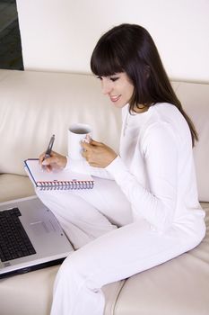 Beautiful latina woman is sitting on a cream color sofa with her computer and a cup of coffee working from home