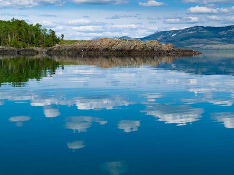 Rocks and green forested hills at Lake Laberge, Yukon Territory, Canada are mirrored on water surface on beautiful summer day.