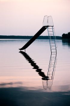 Metal playground slide standing in calm water of big lake.