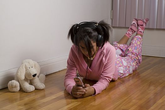 Teenage gir wearing a pink pajama lying on the floor, listing to music and texting on a pink cellphone