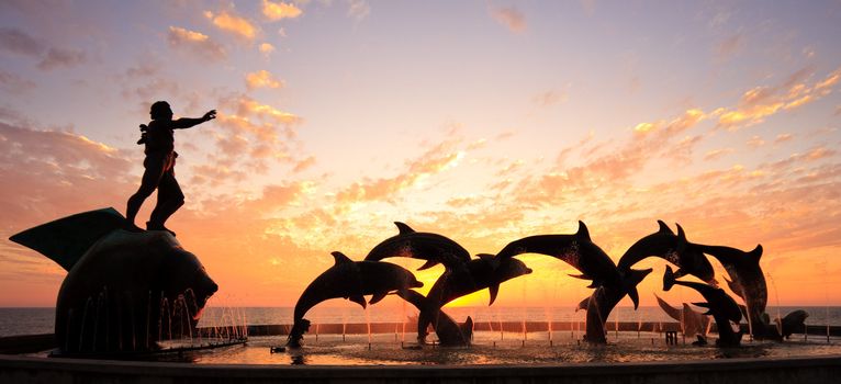 Statues of Dolphins in fountain in front of bright orange sunset over the ocean