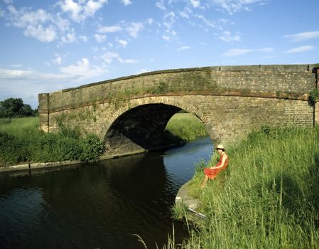 Young lady in red dress sitting on the bank of a picturesque canal by a stone bridge