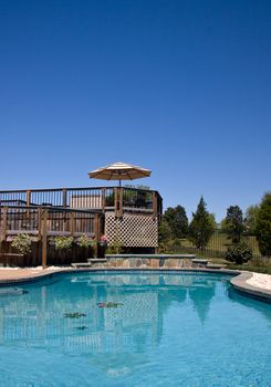 Blue swimming pool looking towards a wooden deck with umbrella and seats