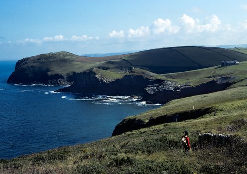 Cornish coast path around rugged headlands with a hiker in red top walking to a stone wall