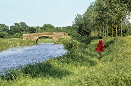 Young lady in red dress walking along a canal towpath towards a stone bridge