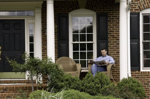 Middle aged man reading a newspaper on the porch of an expensive suburban house