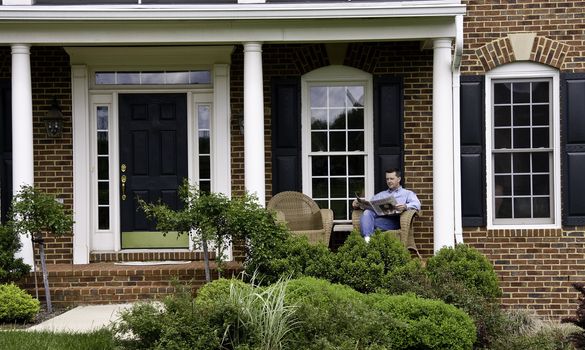 Middle aged man relaxing on the porch of an expensive house reading a newspaper