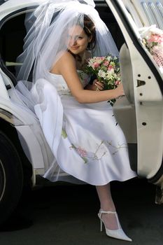 Smiling bride with bouquet exiting wedding car limousine door with sunlight on veil.