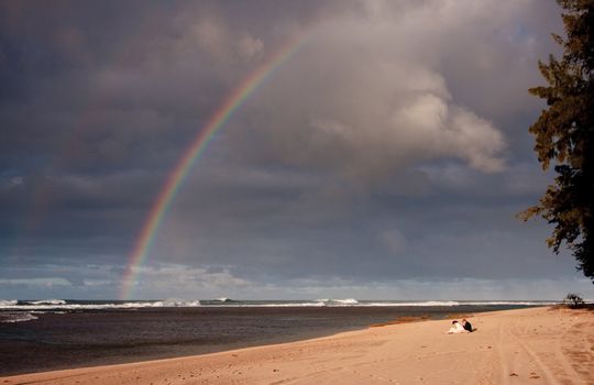 Broad sandy beach with a couple sitting on the sand and viewing a rainbow over the ocean