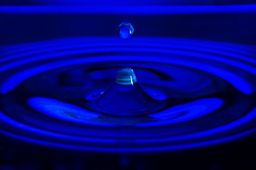 The water drop falls in dark blue water with dispersing circles close up
