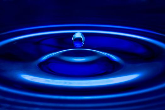 The water drop falls in dark blue water with dispersing circles close up
