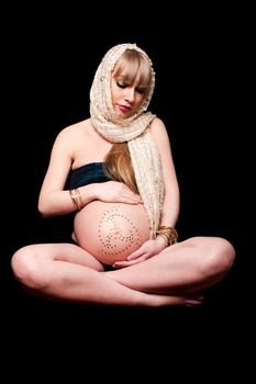 Beautiful pregnant Caucasian woman with head scarf sitting in lotus position holding naked belly covered with rhinestones, isolated