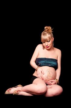 Beautiful pregnant blond girl holding her bare belly covered in rhinestones while looking down and sitting, isolated