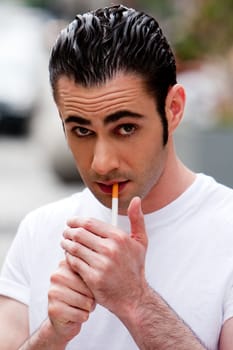 Handsome Caucasian man wearing white shirt lighting a filter cigarette, on a blurred outdoor background