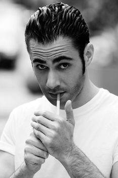 Handsome Caucasian man wearing white shirt lighting a filter cigarette, on a blurred outdoor background