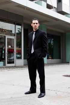 Handsome Caucasian business man in black suit and hands in pocket standing on street in front of a modern building