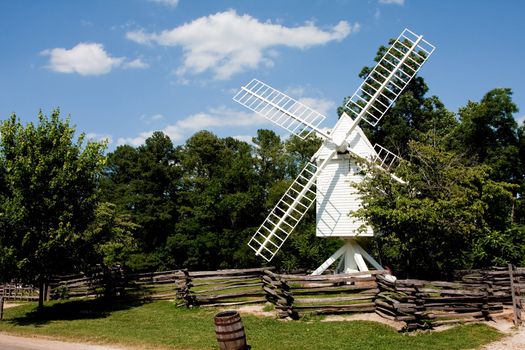 A white wooden windmill in a small meadow behind a wooden fence on a summer day with blue skies surrounded by trees.