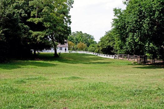 White colonial wooden farm house with a meadow grass field and trees in front barricaded with a white fence.