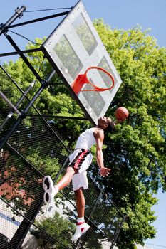 Sporty handsome African-American man dressed in white jumping in the air reaching for the basket while playing basketball in an outdoor court on a summer day trying to dunk the ball