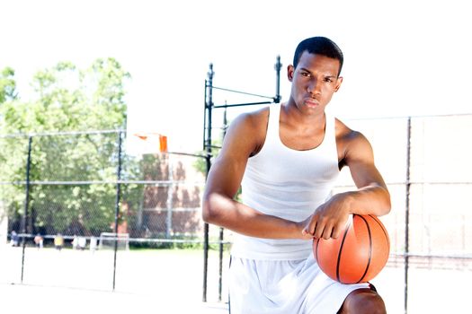 Handsome sporty African-American male basketball player with attitude dressed in white standing with his ball outdoor on a summer day in a basketball court.