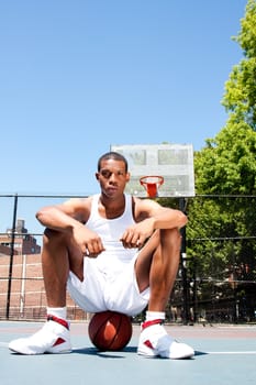 Handsome sporty African-American male basketball player with attitude dressed in white sitting on his ball outdoor on a summer day in a basketball court.