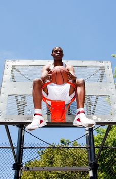 Handsome sporty African-American male basketball player dressed in white and holding his ball outdoor on a summer day in a basketball court while sitting in the hoop.