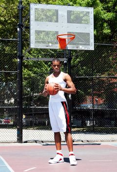 Handsome sporty African-American male basketball player with attitude dressed in white standing holding his ball with both hands outdoor on a summer day in a basketball court.