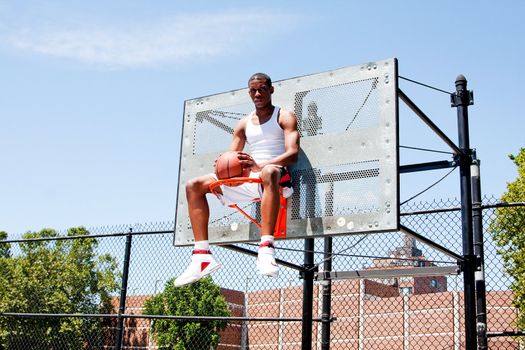 Handsome sporty African-American male basketball player dressed in white and holding his ball outdoor on a summer day in a basketball court while sitting in the hoop.