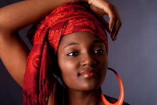 Beautiful African-American woman wearing a traditional tribal red orange head scarf and big orange hoop earrings, with arm over her head, isolated.