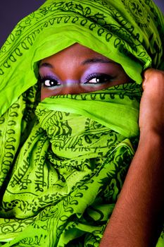 The face of an innocent beautiful young African-American woman covering her mouth showing only her eyes with green headwrap and purple-green makeup, isolated