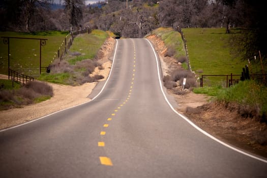 Lonely country road twisting around and over the hill in the distance, fences and green grass on both sides