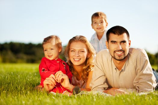 Happy family of 4 people lying ona grass under summer sun. Focus is on the man.