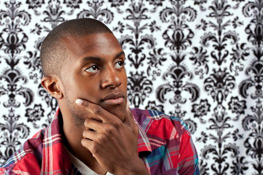 A young African American man in his twenties and his hand on his chin thinking deeply about something in front of a damask style background. Shallow depth of field.