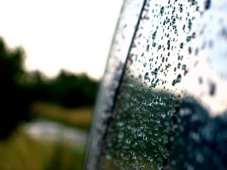 The rain drops on glass of car. Sky and tree in the background.