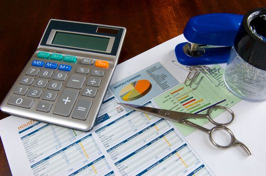 budget spreadsheet on a wooden desk with a calculator, scissors, stapler, and paper clips